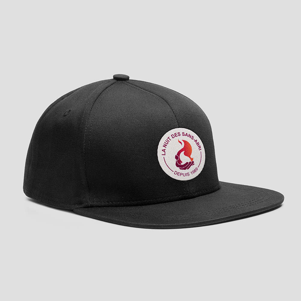 Casquette noire, sur laquuelle est brodée le logo de la Nuit des Sans-Abri. Logo moderne, circulaire, représentant une flamme. Palette de couleurs vives dans les teintes orange et aubergine.